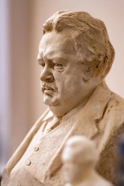 Cream-colored bust of a heavy-set G.K. Chesterton, seen from the chest up, looking left with a pensive expression. He has a prominent mustache and wears a suit jacket over a collared shirt.